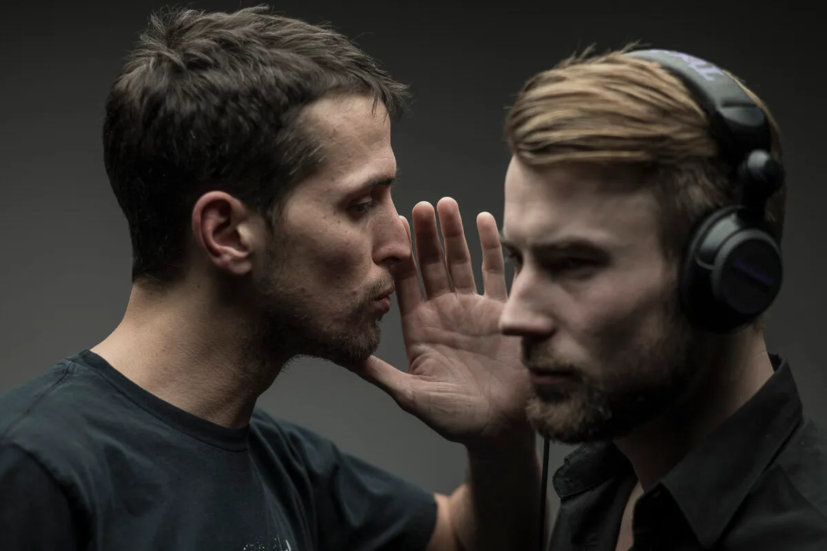 Ben Fein whispering into the ear of a man wearing headphones